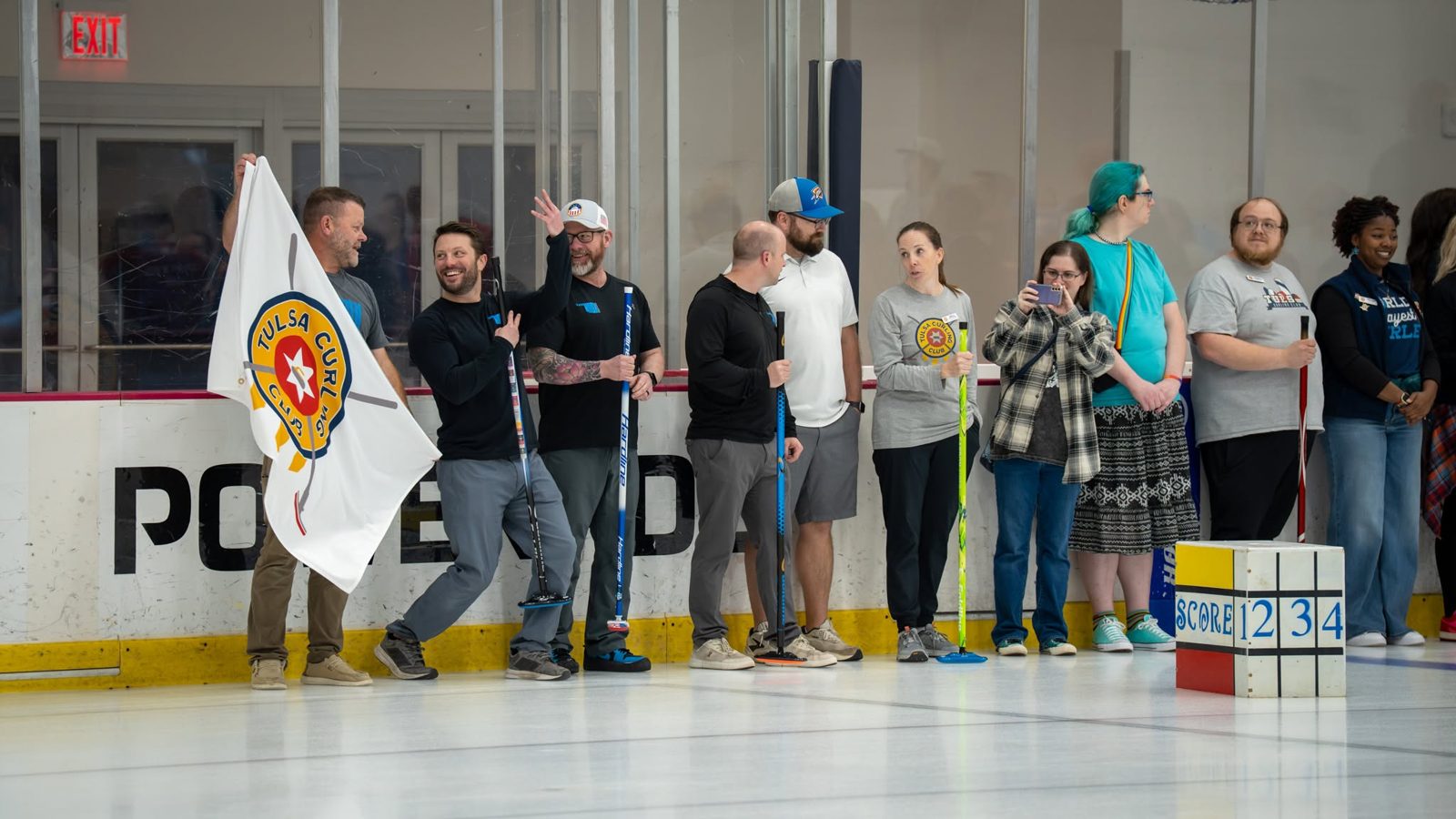 Tulsa Curling Club members line up along the boards with brooms, one waving the club flag